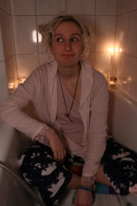 Eine blonde junge Frau die in flauschigen Pyjama Hosen mit Eisbäraufdruck bei Kerzenschein in einer Badewanne sitzt und schmunzelt. Corona Lockdown bedingt ist nicht nur der Aufnahmeort dieses Bildes sondern auch ihr leichter Ansatz vom nachfärben. Das ist Hopia.