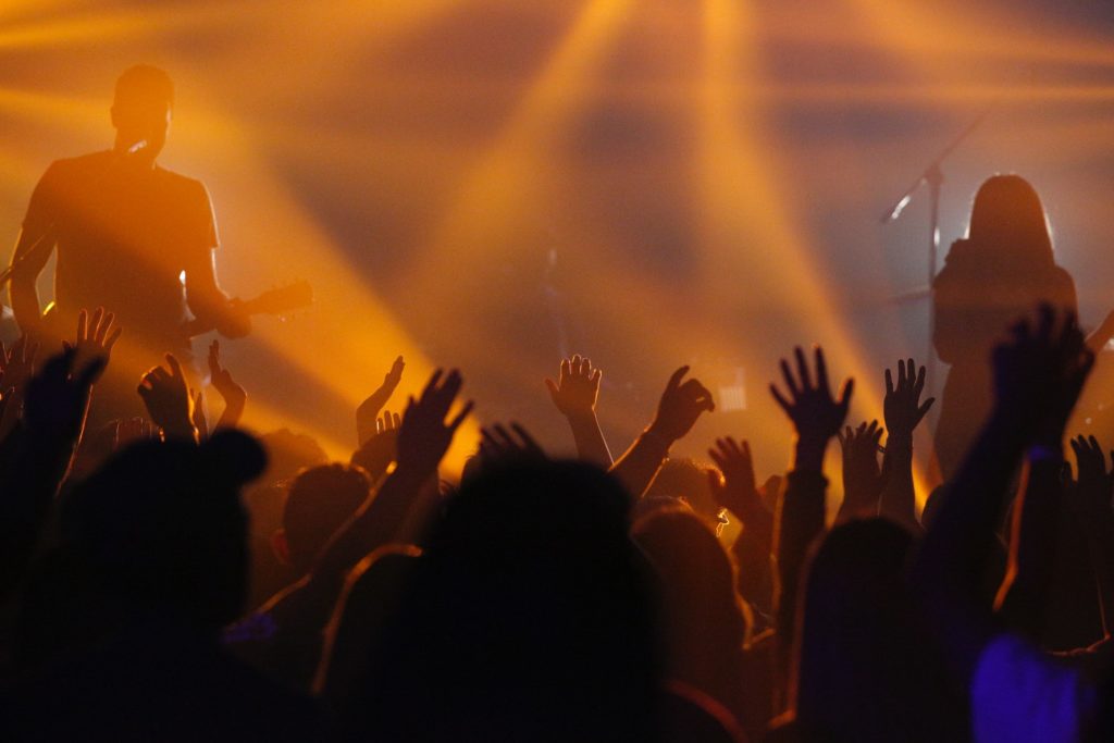 Ein Konzertbild, aufgenommen aus dem Zuschauerraum. Man sieht die Silhouetten des Publikums die ihre Arme nach oben in das orangene Licht strecken