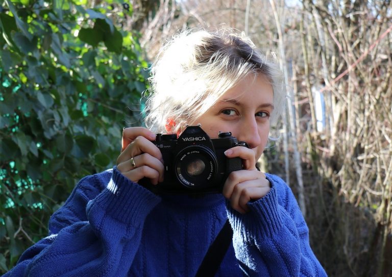 Blonde junge Frau in blauem Pulli schaut hinter einer analogen Kamera hervor